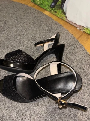 Svarta glittriga skor med klack - Säljer ett par svarta skor med hög klack och öppen tå. Främre delen har glittrigt material och remmen runt ankeln har ett snyggt spänne i guld. Snyggt lackad finish på klacken och remmarna. Perfekt för dig som gillar att sticka ut med lite extra bling. Storlek 39. De är helt nya 