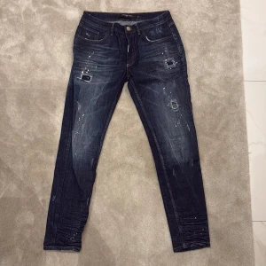 Mörkblå jeans med slitningar från Alessandro Zavetti - Säljer ett par mörkblå jeans från Alessandro Zavetti med coola slitningar och färgstänk. Byxorna har klassisk femficksdesign, knappgylf och raka ben. Perfekta för dig som vill ha en trendig och avslappnad stil.