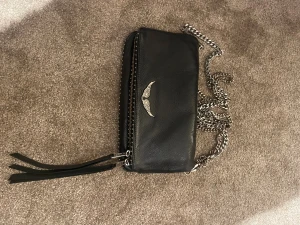 Svart väska från Zadig & Voltaire - Säljer en svart väska i skinn från Zadig & Voltaire med silverfärgad kedjerem och dragkedja. Väskan har en ikonisk vingdetalj framtill och dekorativa läderband vid dragkedjan. Väskan är köpt här på Plick för 2 år sedan och är i bra skick! Kedjan har dock blivit lite rostig men inget man lägger märke till.