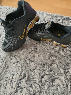 Nike Shox svarta sneakers med guld - Nike Shox sneakers i svart med guldiga detaljer och metallicfärgad Shox-plattform. Snyggt broderad guld Swoosh på sidan, ventilerande mesh och perforerat syntetmaterial. Skon har snörning och en unik design med fjädrande sula för extra komfort.