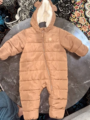 Brun pufferoverall med öron från H&M - Säljer en brun pufferoverall från H&M i storlek 74 med mysig vit fleecefoder. Overallen har huva med söta öron, hel dragkedja och quiltad design. Perfekt för kalla dagar och håller barnet varmt och bekvämt.
