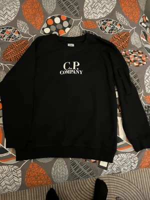 Svart sweatshirt från C.P. Company - Svart sweatshirt från C.P. Company med vit logotyp på bröstet och en ikonisk linsdetalj på ärmen. Tröjan har rund hals, ribbade muddar och är långärmad. Perfekt för dig som gillar streetwear och vill ha en clean look. • Storlek: Märkt M men passar som en S. 