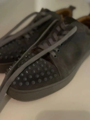 Christian Louboutin svarta sneakers med nitar - 44/43Svarta sneakers från Christian Louboutin med ikoniska röda sulor och coola nitar på tån. Skorna har snörning och är gjorda i mocka med insida i brunt skinn. Perfekt för dig som vill sticka ut med en lyxig och edgy stil.