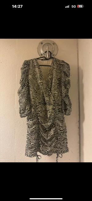 Leopardmönstrad kortklänning med snörning - Säljer en kort klänning med leopardmönster i beige och svart. Klänningen har v-ringad omlottfront, puffiga trekvartsärmar och snygg rynkning med snörning längs sidorna. Perfekt för dig som gillar att sticka ut med djurmönster och coola detaljer.