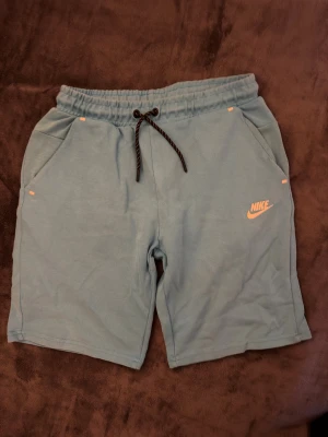Nike ljusblå shorts med snörning - Nike shorts i ljusblått med elastisk midja och svartvit snörning. Mjukt bomullsmaterial, två fickor fram och en bakficka med dragkedja. Orange Nike-logga framtill ger en sportig vibe. Perfekta för chill dagar eller träning.