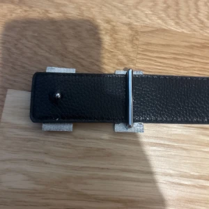 Mörkblått dubbelsidigt skärp med H-spänne - Säljer ett stilrent marinblått skärp med ikoniskt H-spänne i silverfärgad metall. Bältet är gjort i präglat skinn med vita kontrastsömmar och klassisk design. Perfekt för dig som vill ha en lyxig accessoar med statement-känsla.