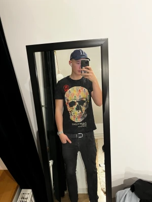 Philipp Plein T-shirt  - Säljer en svart t-shirt från Philipp Plein med ett stort färgglatt döskalle-tryck på bröstet och textdetaljer. T-shirten har rund hals och korta ärmar, perfekt för dig som gillar statement-plagg och exklusiv design.
