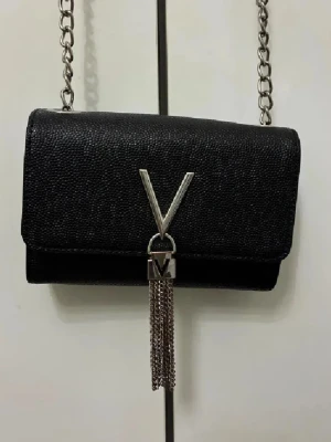 Svart axelväska från Valentino - Snygg svart axelväska från Valentino med silverkedja och V-logga framtill. Väskan har en stilren, rektangulär form och är tillverkad i skinn med en lätt textur. Framtill hänger dekorativa silverfransar från loggan för extra edge.