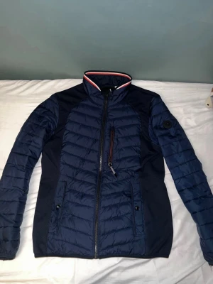 Mörkblå pufferjacka från Tom Tailor - Snygg mörkblå pufferjacka från Tom Tailor med quiltad design och hög krage med rödvit detalj. Jackan har dragkedja framtill, bröstficka med dragkedja och två sidofickor med knappar. Perfekt för kyliga dagar och har en sportig vibe.