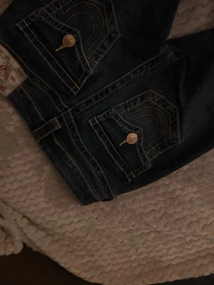 True Religion mörkblå jeans med kontrastsömmar - Säljer ett par mörkblå jeans från True Religion med ikoniska kontrastsömmar och stora fickor bak med guldfärgade knappar. Jeansen har klassisk passform och är tillverkade i slitstarkt denim. Snygga detaljer och logga på knapparna ger en unik look. Älskar dem jätte mycket men har tyvärr blivit för korta för mig.❤️❤️Kostade 1300kr när jag köpte dem.