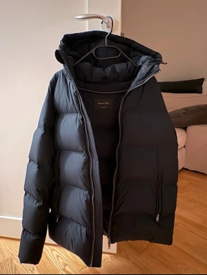 Svart pufferjacka från Massimo Dutti - Svart pufferjacka från Massimo Dutti med huva och två sidofickor med dragkedja. Jackan har en stilren look med quiltad design och rejäla dragkedjor framtill. Perfekt för kalla dagar och har en modern, clean vibe. Knappt använd, inga tecken på slitage eller skavanker. 