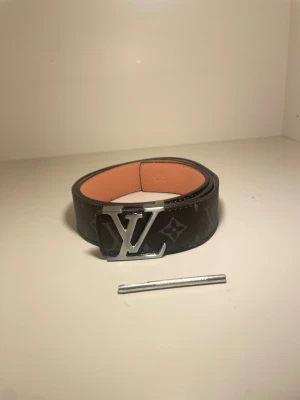 Designer bälte  - Snyggt och stilrent designerbälte i svart monogrammönster med elegant silverfärgat spänne. Bältet är i fint skick och passar perfekt för dig som vill lyfta vilken outfit som helst – både casual och mer uppklätt.  ✔ Klassisk monogramdesign ✔ Silverfärgat spänne ✔ Justerbar längd ✔ Levereras med medföljande pinne för anpassning av storlek ✔ Insida i ljus, mjuk läder-look