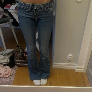 Blå bootcut jeans  - Säljer ett par blå bootcut jeans med snygga broderade detaljer på bakfickorna. Jeansen har låg midja och klassisk femficksdesign. Materialet är jeans och passformen är normal med lite utsvängda ben. Perfekt för dig som gillar detaljer och en cool stil.