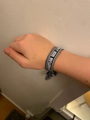 Svart och vit Christian Dior J'Adior armband - Snyggt armband från Christian Dior med texten 'J'Adior' broderat i vitt på svart bakgrund. Armbandet har dekorativa tofsar och knytning i ändarna, samt ett subtilt mönster längs kanterna. Perfekt accessoar för att lyfta din outfit.