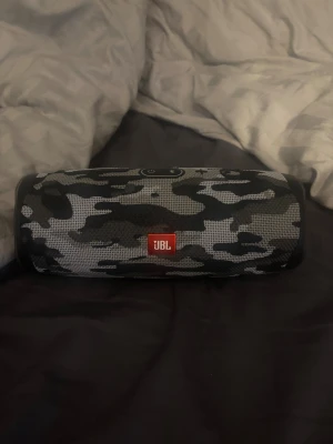 JBL Charge 4 Camouflage - JBL Charge 4 portabel Bluetooth-högtalare i snyggt camouflagemönster. Kraftfullt ljud, lång batteritid och robust design. Högtalaren ser ut att vara i gott skick utan synligt slitage eller defekter. Perfekt för både inomhus och utomhusbruk.