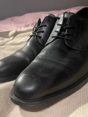 Svarta klassiska skinnskor med snörning - Snygga svarta skinnskor med rund tå och platt sula. Skorna har klassisk snörning och stilren design som passar till både kostym och mer avslappnade outfits. Materialet är slätt skinn och de har en enkel, tidlös look som funkar året runt.