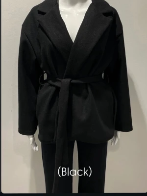 Svart kappa  - Stilren svart kappa med omlottknäppning och bälte i midjan. Jackan har klassisk krage, raka långa ärmar och en rak passform. Materialet känns mjukt och värmande, perfekt för kyligare dagar. Använda ett fåtal gånger. 