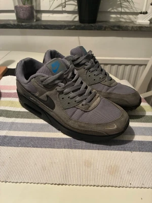 Nike Air Max 90 grå sneakers - Nike Air Max 90 sneakers i grått med svarta detaljer och blå logga. Ovandel i mesh och mocka, klassisk Air Max-sula med synligt luftfönster. Rund tå, snörning och platt sula. Perfekt för dig som gillar stilrena och sportiga sneakers.