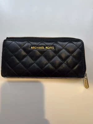 Svart quiltad plånbok Michael Kors - Stilren svart plånbok från Michael Kors i quiltat skinn med guldfärgade detaljer och dragkedja. Plånboken har flera fack för kort och mynt, samt logga i guld på framsidan. Perfekt för dig som gillar lyxig och klassisk design.