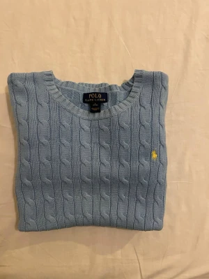 Ljusblå kabelstickad tröja Polo Ralph Lauren - Polo Ralph Lauren Cable-Knit Tröja i en vacker ljusblå färg. Klassisk krage, flätstickad design och den ikoniska gula ponnyn. Storlek Barn L (14-16), passar även vuxen XS/S. Tidlös och stilren! 