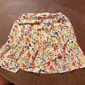 Färgglad blommig omlottkjol - Superfin kort omlottkjol från Name It med färgglatt blommigt mönster i rosa, gul, lila och orange. Kjolen har volangdetalj och resår i midjan.