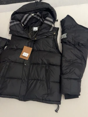 Svart pufferjacka från Burberry - Säljer en svart pufferjacka från Burberry. Jackan har dragkedja, knappar och två fickor med dragkedja framtill. Snyggt Burberry London England-tryck på ärmen. Jätte fin passform. 