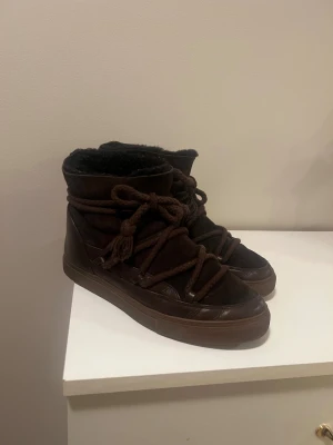 Bruna Inuikii  - Säljer ett par bruna Inuikii boots med snörning och rund tå. Skorna är tillverkade i en mix av mocka och skinn, med en mysig fodrad insida. Perfekta för kalla dagar och har en chunky platt sula för extra komfort. Liten defekt på sista bilden men annars i bra skick 