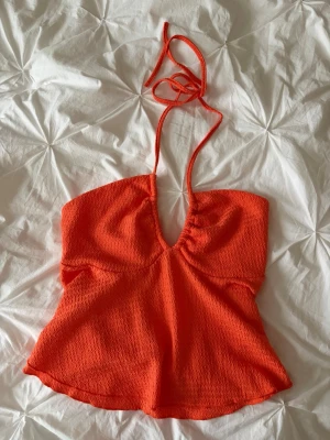Halterneck topp - Säljer en snygg orange topp med knytband i nacken och djup v-ringning. Toppen har en vågig textur i tyget och är lätt utsvängd nedtill. 