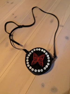 Fin rund axelväska  - Fin axelväska med broderad röd fjäril på framsidan och sydda vita knappar. Köpt second hand men i extremt bra skick. Lite retro/vintage vibbar?