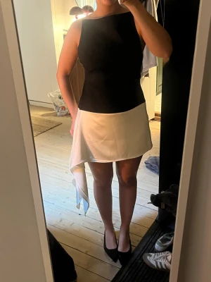 Svartvit kortklänning med asymmetrisk detalj - Säljer en svartvit kortklänning med cool asymmetrisk volangdetalj på kjolen. Klänningen har ärmlös design och rak halsringning, med svart överdel och vit kjol i mjukt, slätt material. Slutsåld och helt oanvänd med prislapp kvar.