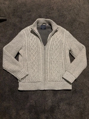Fodrad cardigan - Snygg grå stickad kofta med dragkedja och mysigt fleecefoder på insidan. Koftan har ett klassiskt flätmönster framtill, ribbade muddar och hög krage. Perfekt för kyliga dagar och enkel att matcha med jeans eller chinos.