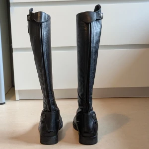 Svarta ridstövlar från CRW i skinn - Säljer ett par svarta ridstövlar från CRW i äkta skinn. Stövlarna har snörning framtill, rund tå och platt sula. De är höga med dragkedja baktill och dekorativa knappar upptill. Perfekta för dig som gillar ridning och vill ha en klassisk look. Två små defekter på utsidan av ridstövlarna, visas på bild.