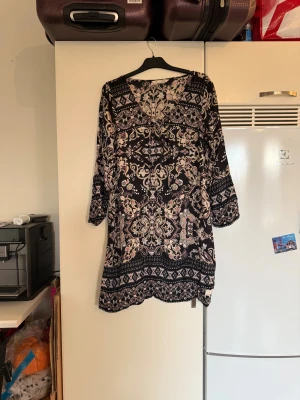 Svart kortklänning med bohemiskt mönster - Säljer en svart kortklänning med bohemiskt mönster i beige, rosa och vitt. Klänningen har långa ärmar, v-ringning med snörning och ett detaljerat print med blommor och fjärilar. Perfekt för dig som gillar unika prints och avslappnad stil. Från märket Odd Molly. Storlek 3 som enligt deras storleksguide motsvarar en L. Mycket fint skick, jag hittar inga synliga anmärkningar eller defekter. 