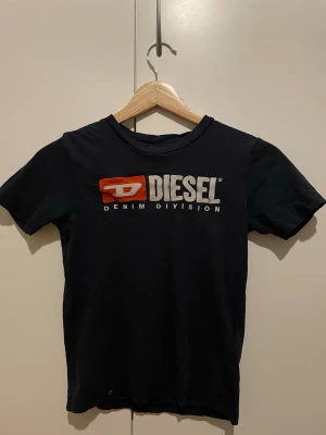 Svart t-shirt från Diesel - Svart t-shirt från Diesel med klassisk logga i vitt och rött framtill. Modellen har rund hals och korta ärmar. Materialet är mjuk bomull som känns skön mot huden. Perfekt för dig som gillar streetwear och vill ha en enkel men snygg look.