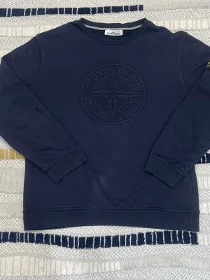 Mörkblå sweatshirt från Stone Island - Snygg mörkblå sweatshirt från Stone Island med stort broderat logomärke på bröstet och klassisk patch på vänster ärm. Tröjan har rund hals, långa ärmar och är gjord i mjuk bomull. Perfekt för dig som gillar streetwear och vill ha en clean look.