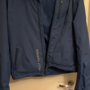 Blå vindjacka från Helly Hansen - Snygg marinblå vindjacka från Helly Hansen med dragkedja framtill, två sidofickor med dragkedja och logga på bröstet och längs sidan. Jackan är tillverkad i polyester och har en klassisk, sportig look med lång ärm och normal passform. Helt ny nästan 