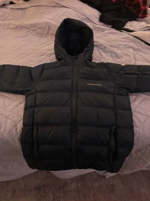 Svart Peak Performance pufferjacka - Svart pufferjacka från Peak Performance med huva och justerbara ärmslut med kardborre. Jackan har en quiltad design och dragkedja framtill. Perfekt för kalla dagar och har en clean, sportig look som funkar till det mesta. Den har ett hål som jag har täckt men d syns knappast