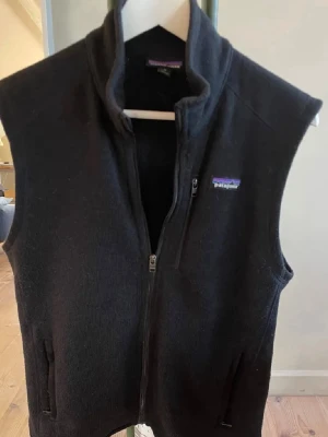 Svart fleeceväst från Patagonia - Svart fleeceväst från Patagonia med hög krage och hel dragkedja framtill. Västen har två sidofickor med dragkedja och ett litet broderat Patagonia-märke på bröstet. Perfekt för lager-på-lager och chill stil.