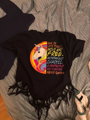 Svart t-shirt med enhörning och fransar - Cool svart t-shirt med färgglad enhörning och texttryck på framsidan. T-shirten har korta ärmar och fransar längst ner för en extra edgy look. Perfekt för dig som gillar unika och färgstarka plagg.