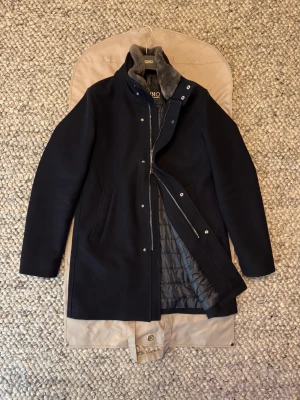 Herno rock  - Snygg mörkblå rock från Herno med klassisk rak passform, quiltat innerfoder och mjuk krage i bäverpäls. Jackan har diskreta sidfickor, dold dragkedja och knappar framtill. Perfekt för kyliga dagar när du vill hålla stilen.