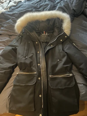 Svart dunjacka med fuskpäls från Horspist - Svart dunjacka från Horspist med fluffig fuskpäls runt huvan, flera dragkedjor och stora fickor framtill. Jackan har en snygg badge på ärmen och quiltad insida. Perfekt för kalla dagar när du vill hålla stilen.