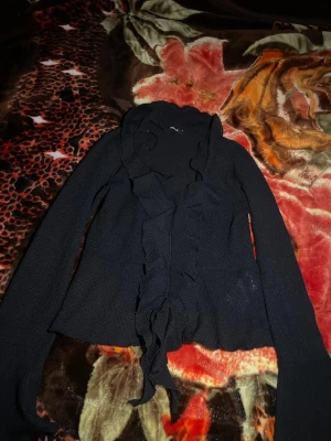 Svart kofta med volangdetaljer - Snygg svart kofta med långa ärmar och volang längs hela framkanten. Materialet känns mjukt och har en lätt struktur, perfekt för dig som gillar en stilren look med lite extra edge. Passar dig som vill ha något enkelt men ändå med coola detaljer.