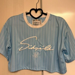 Blå croppad topp från SikSilk - Säljer en ljusblå croppad topp från SikSilk med vita vertikala ränder och broderad logga framtill. Toppen har vit krage och är kortärmad, tillverkad i ett mjukt syntetmaterial som känns sportigt och fräscht. Perfekt för dig som gillar streetwear och snygga detaljer.