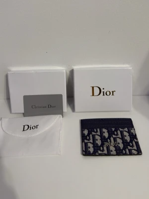 Blå Dior plånbok med mönster - Supersnygg plånbok från Dior i marinblått och beige med ikoniskt Dior-mönster. Slimmad design med plats för kort, perfekt att ha i fickan eller väskan. Kommer med originalförpackning och dustbag. Metallogo framtill ger extra lyxig känsla.