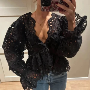 Svart broderad blus med knyt - Säljer en svart blus med broderade detaljer och v-ringning. Blusen har långa puffärmar, knyt i midjan och spetskantade avslut. Materialet är lätt och genombrutet vilket ger en cool och unik look. 
