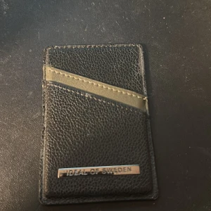 Svart plånbok från iDeal of Sweden - Slimmad svart plånbok från iDeal of Sweden med stilren design och metallogo framtill. Plånboken har två snedställda kortfack och är tillverkad i skinn med en lätt textur. Perfekt för dig som vill ha ett minimalistiskt och snyggt kortfodral.