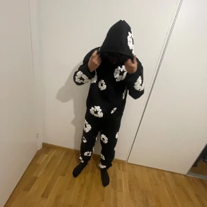 Svart hoodie med vita blommor - Svart hoodie med stora vita blommönster över hela tröjan och huvan. Tröjan har en rymlig passform, långärmade ärmar och en klassisk huva. Perfekt för dig som gillar streetwear och vill sticka ut med ett coolt mönster.