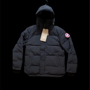 Svart dunjacka från Canada Goose - Säljer en svart dunjacka från Canada Goose med huva och klassisk logotyp på vänster ärm. Jackan har quiltad design, dragkedja framtill och ribbade muddar. Perfekt för kalla dagar och har en clean, stilren look som funkar till det mesta.