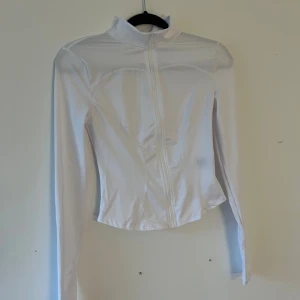 Vit croppad träningsjacka med dragkedja - Snygg vit träningsjacka med croppad passform och hög krage. Jackan har lång ärm och hel dragkedja framtill. Materialet är lätt och syntetiskt, perfekt för en sportig look. Enkel design utan mönster eller extra detaljer.