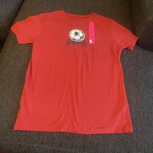 Röd t-shirt med fotbollstryck FSFC - Röd t-shirt från FSFC med tryck av en fotboll och text på bröstet. Klassisk rund hals och korta ärmar. Perfekt för dig som gillar fotboll och vill ha en enkel, sportig stil. Gjord i mjuk bomull för skön känsla hela dagen.